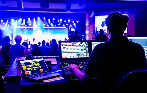 Live event production technician managing audio, video, and lighting systems from a control booth during a stage performance.