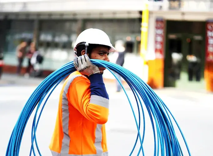 Construction worker carrying blue cables wearing safety gear on a city street.